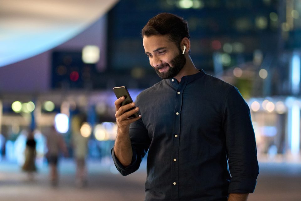 Rich successful indian business man professional entrepreneur wearing earbud standing outdoors on night city street holding cell phone using smartphone watching video or having mobile chat call.
